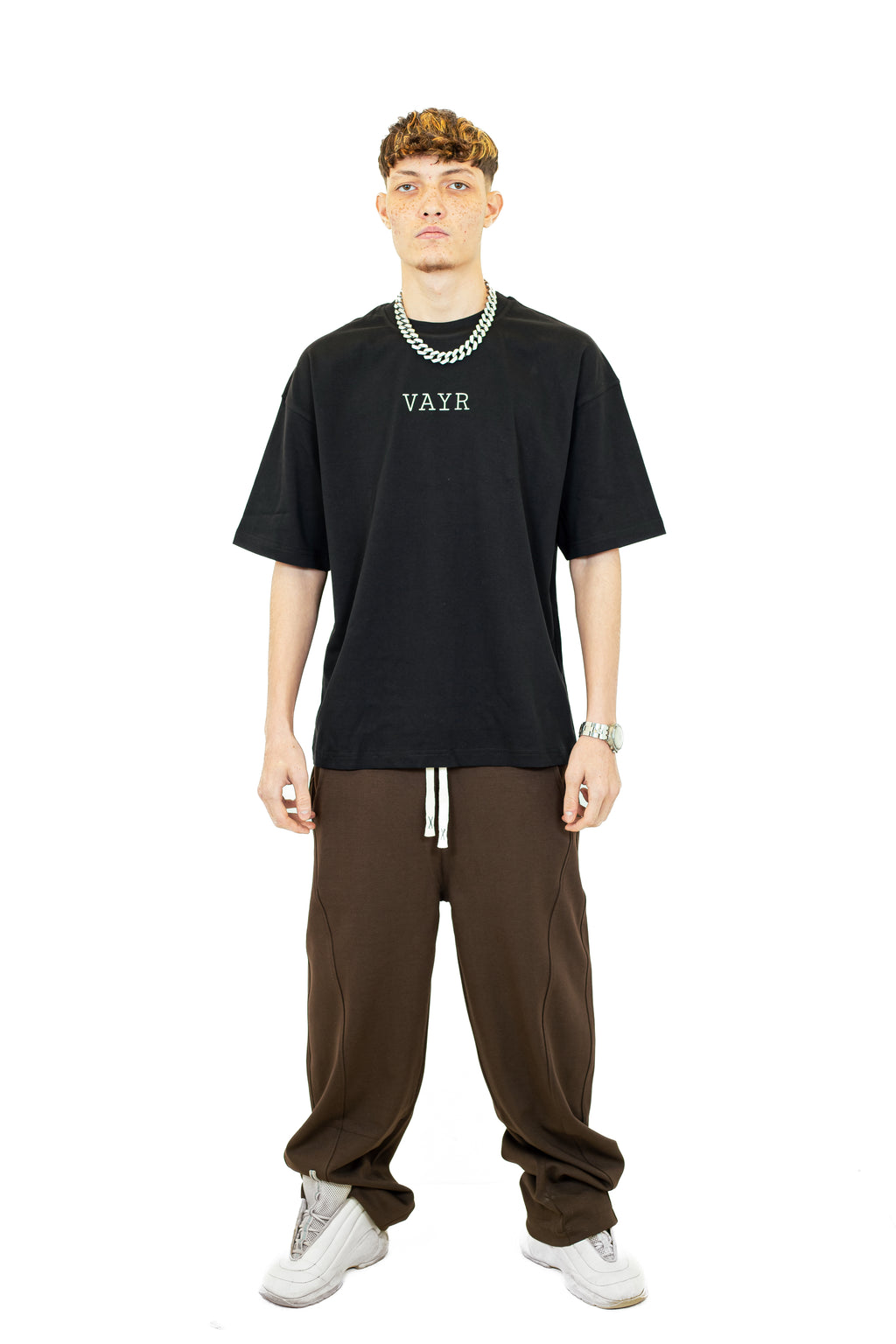 Man wearing a black t-shirt with 'VAYR' logo, brown pants, and white sneakers on a white background