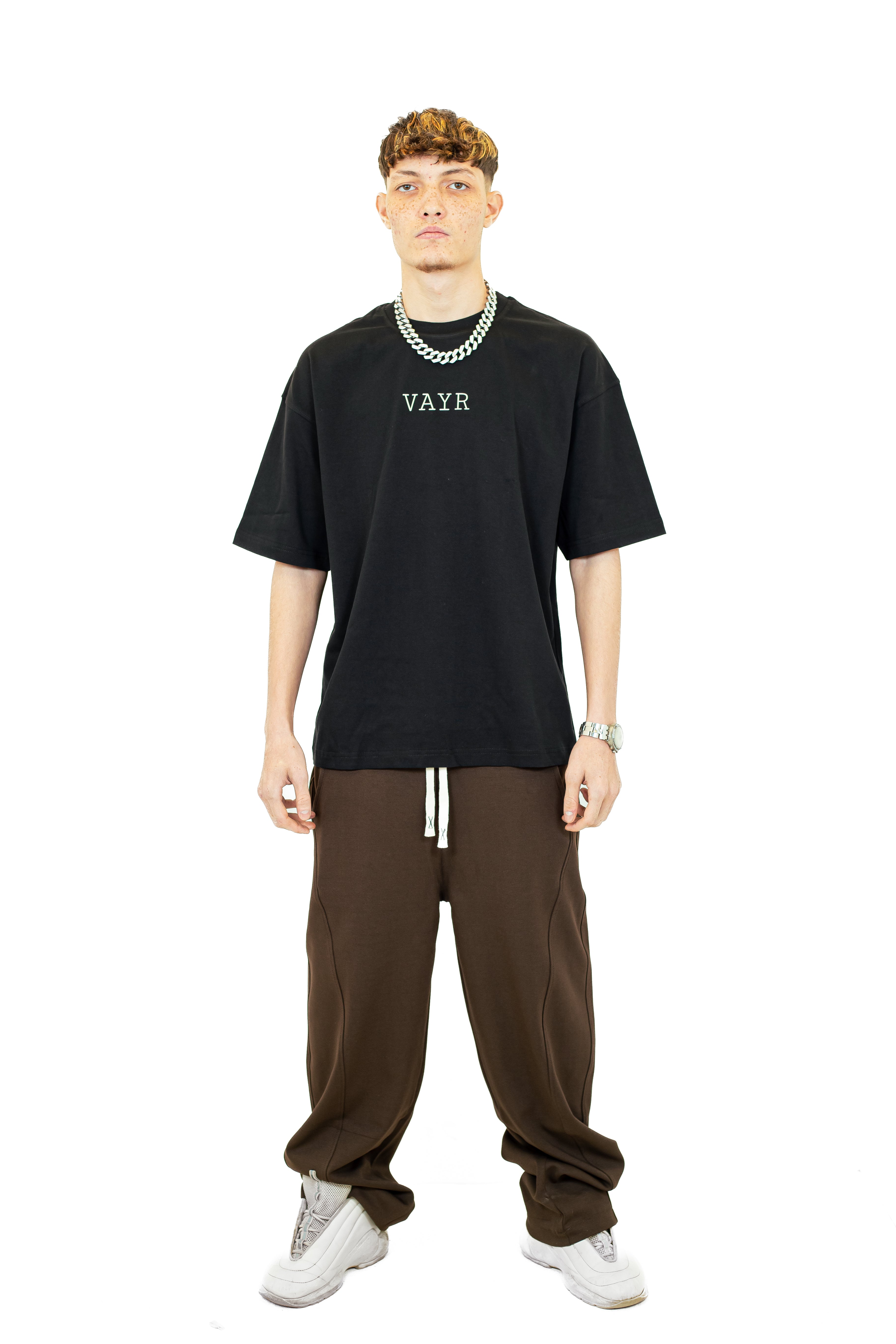Man wearing a black t-shirt with 'VAYR' logo, brown pants, and white sneakers on a white background