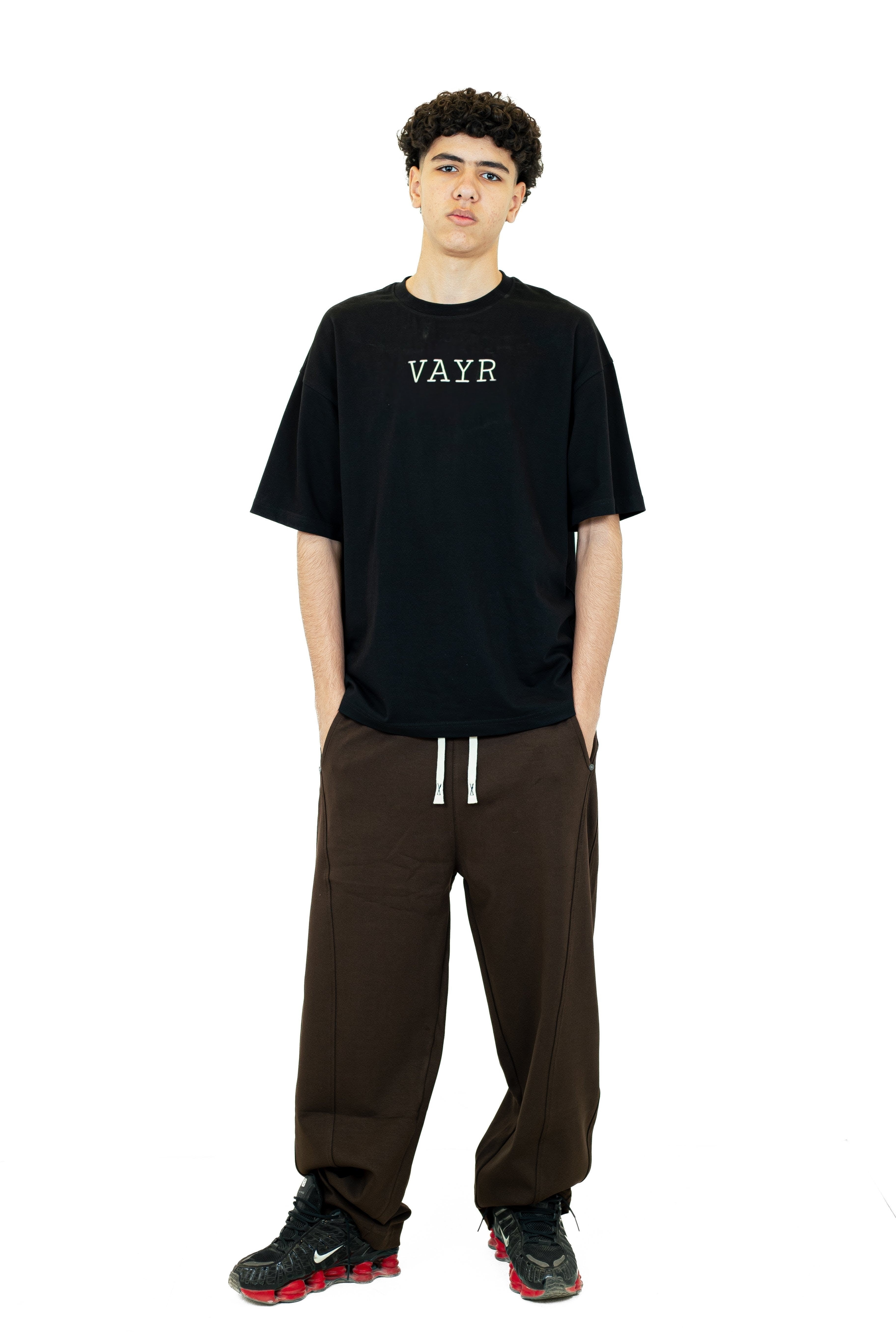 Person wearing a black t-shirt with 'VAYR' logo and brown pants on a white background